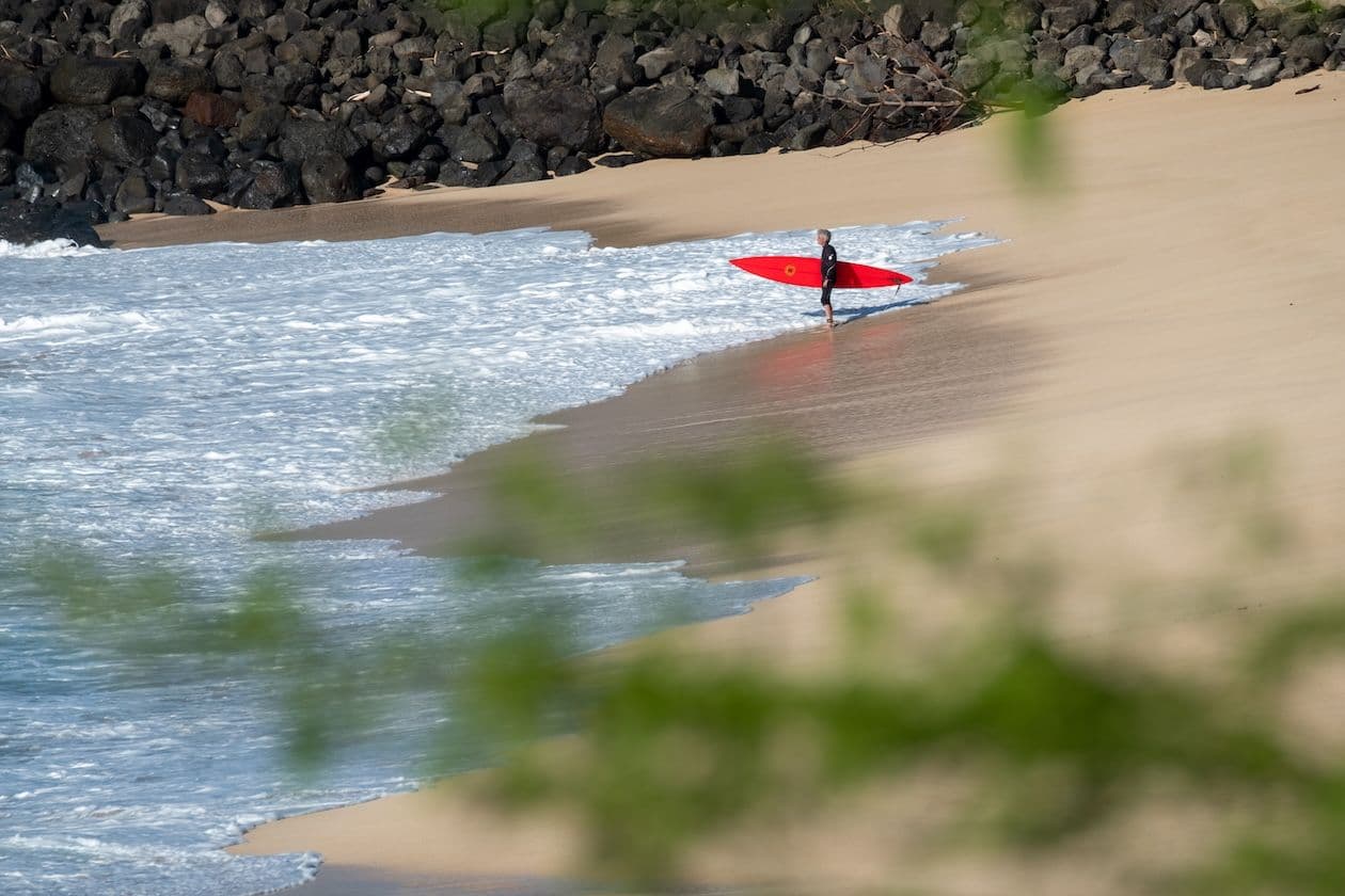 Man with surfboard getting into the ocean in front of large rock wall. Oahu.