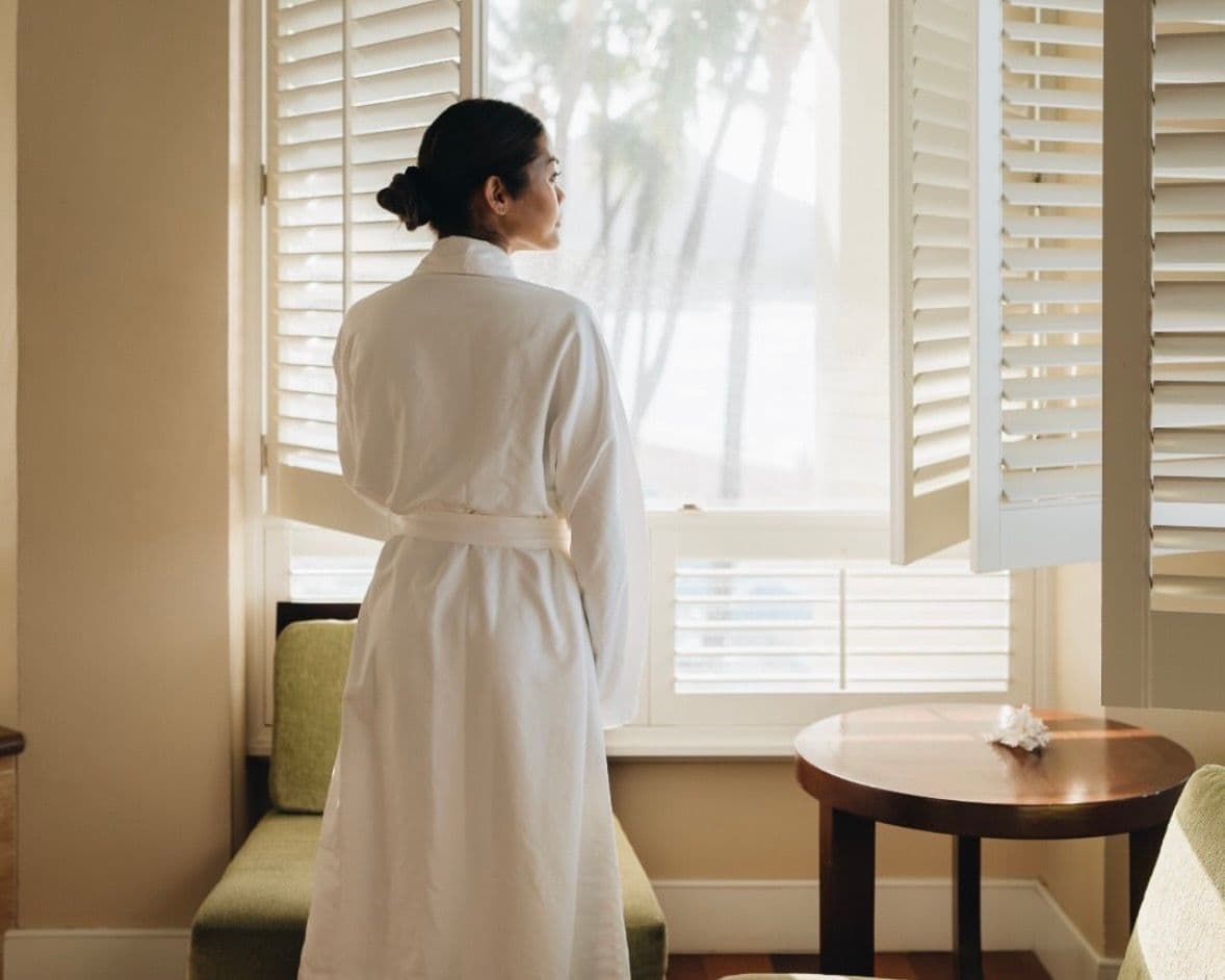 Woman standing at a window in a white robe.